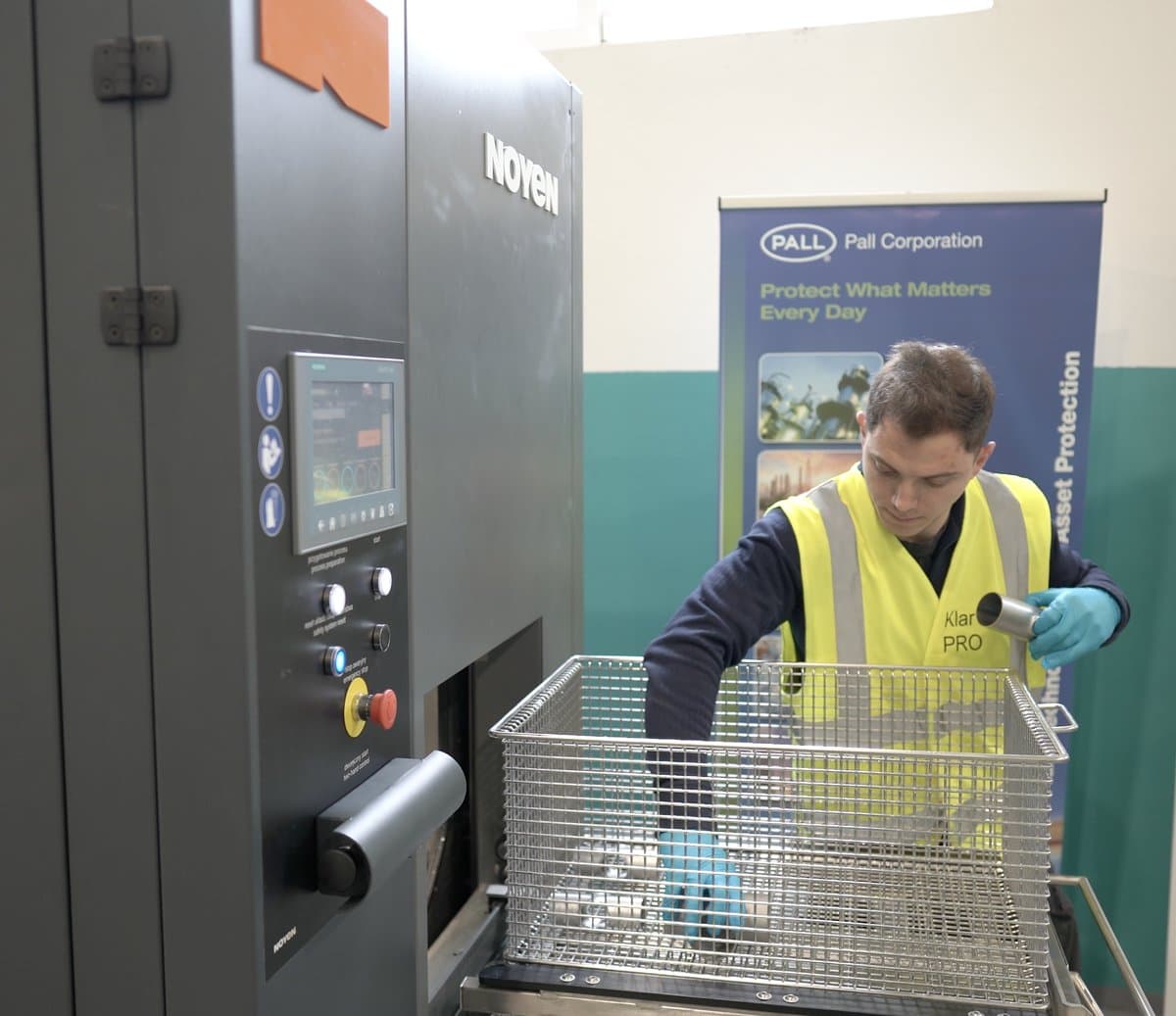 Klarwin operator loading a parts basket into a Noyen automated washing machine next to a Pall Asset Protection banner