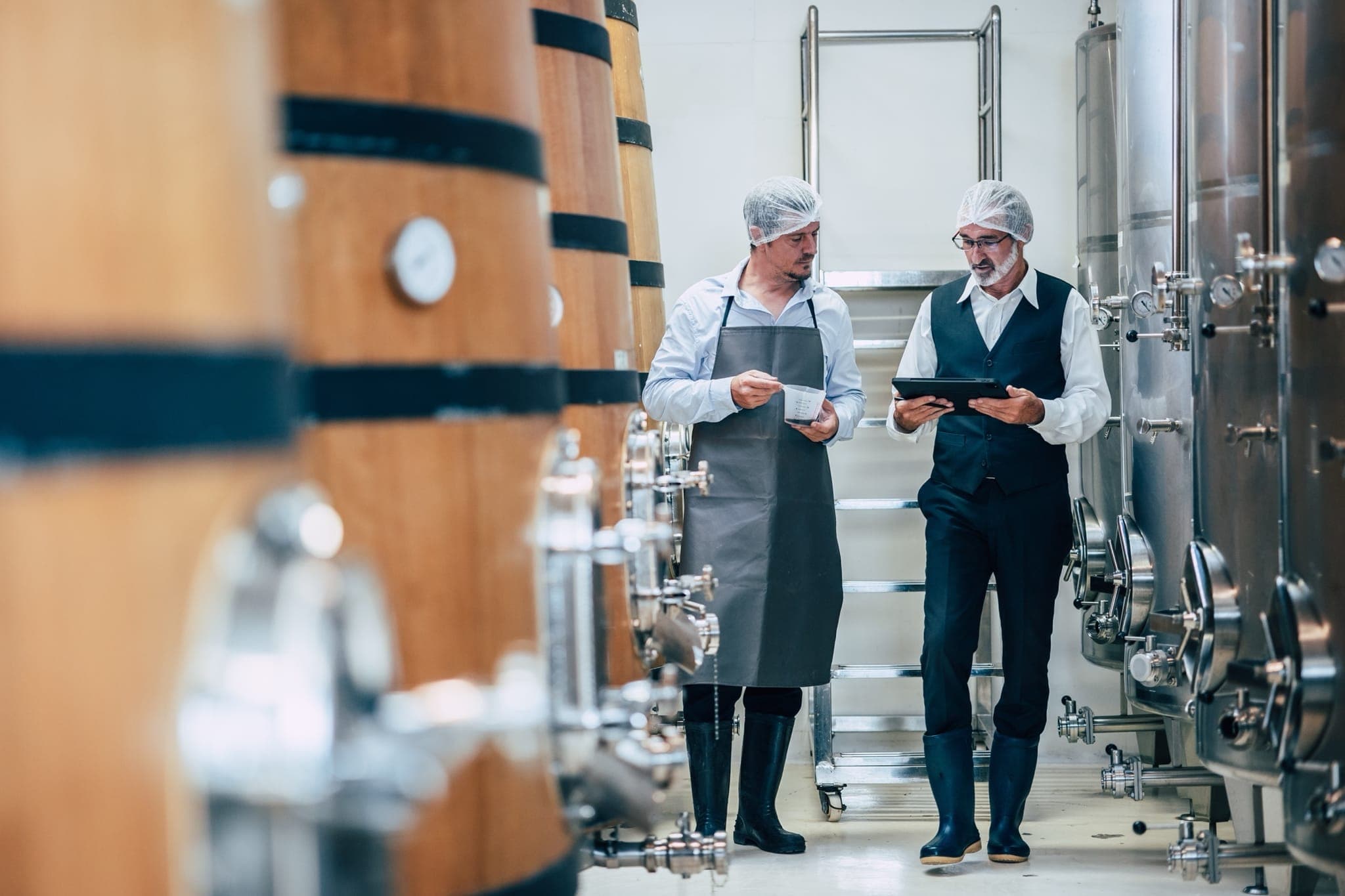 Winemakers checking production data on a tablet between oak barrels and stainless steel tanks